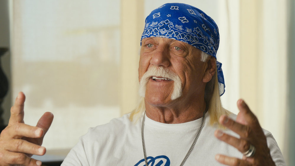 Hulk Hogan in a white shirt and blue bandanna holding his hands up