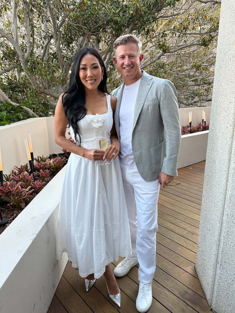 Katie Ginella in a white dress, holding a flute of champagne and posing with Matt Ginella, who is wearing all white and a grey blazer