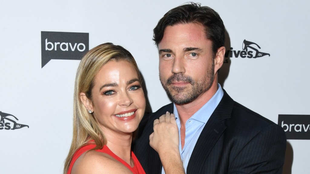Denise Richards in a red dress leaning on Aaron Phypers, who is waring a dark blue suit