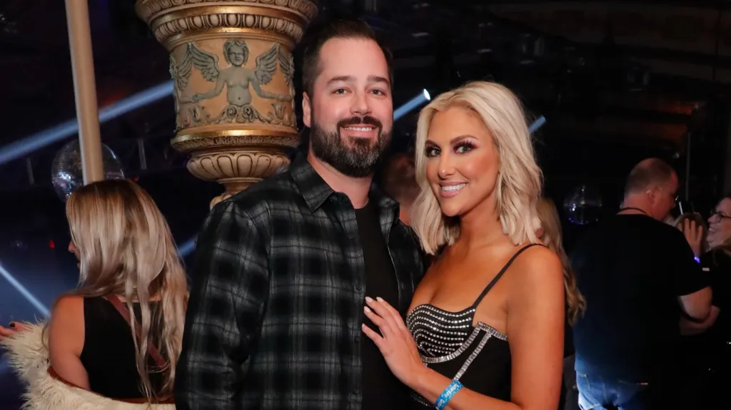 Travis Mullen and Gina Kirschenheiter backstage at BravoCon 2022, Gina is posing with her hand on Travis's chest