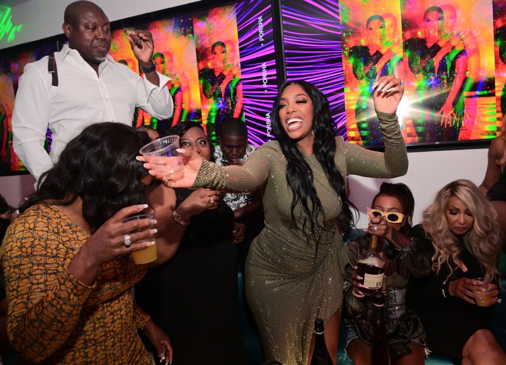 Porsha Williams in a green dress, holding out a cup in the middle of a club, while Simon Guobadia watches from the back
