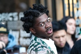 Lil Nas X in a green coat and gold glasses, tilting his head and posing in front of a crowd of people