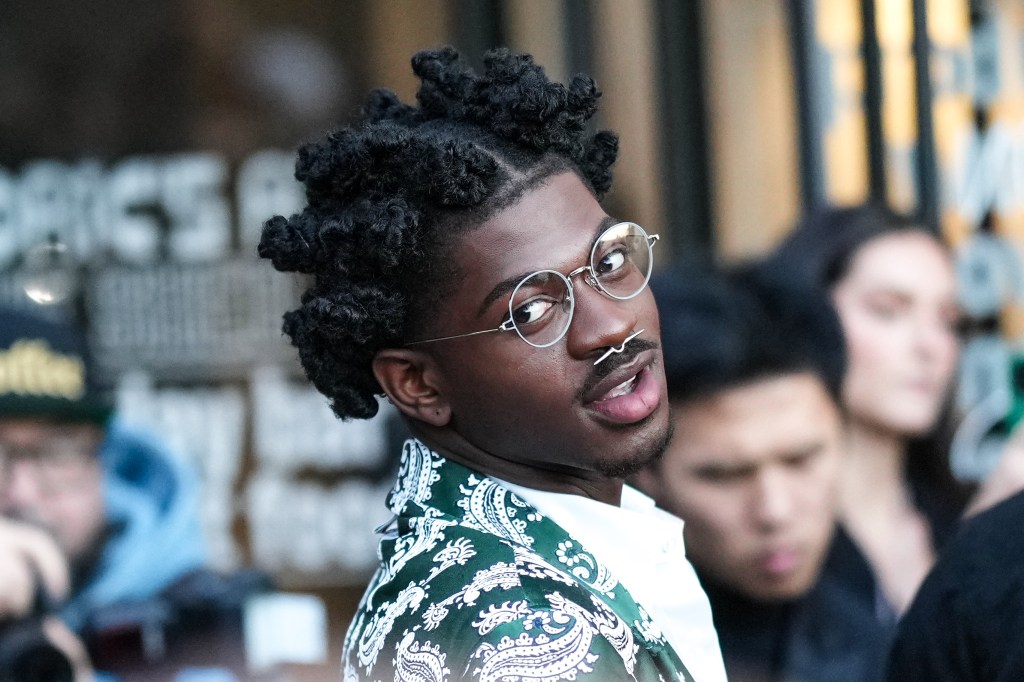 Lil Nas X in a green coat and gold glasses, tilting his head and posing in front of a crowd of people