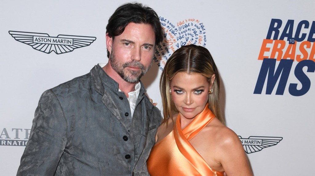 Aaron Phypers in a grey suit posing with Denise Richards, who is wearing an orange dress and looking down