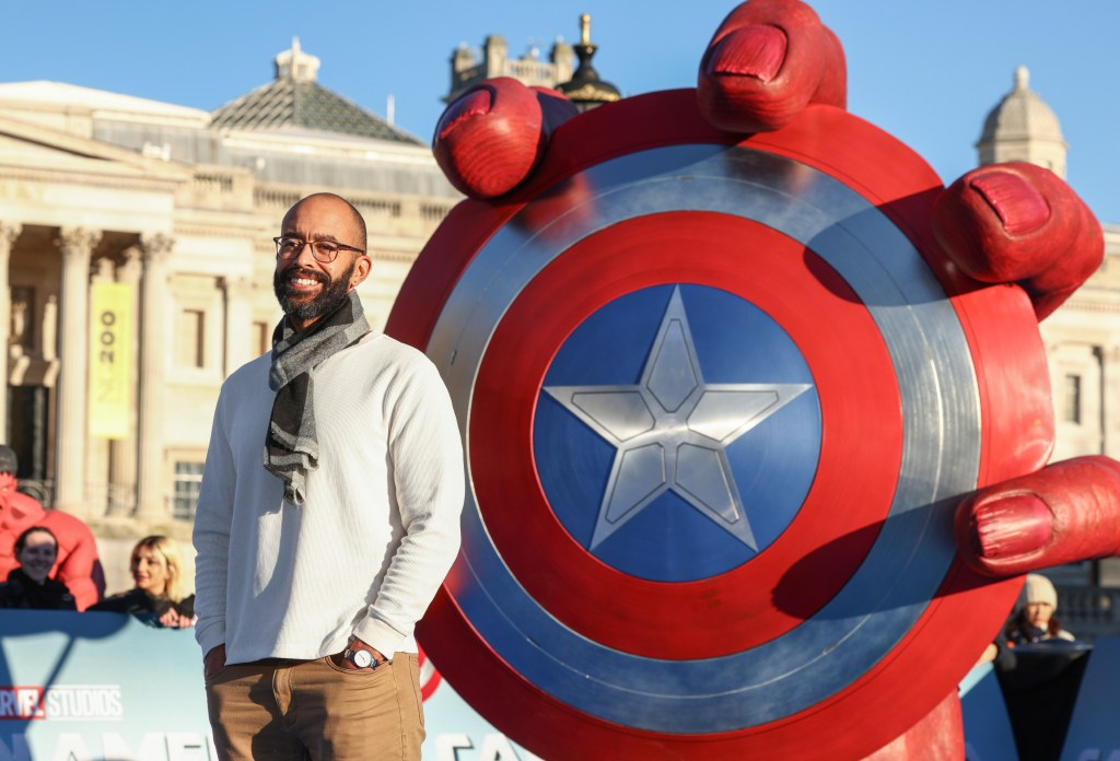 Nate Moore in a white sweater posing in front of a statue of Red Hulk holding a Captain America shield