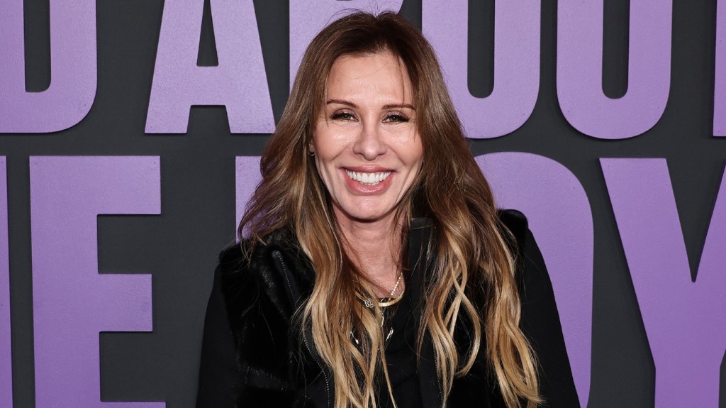 Carole Radzwill in a black coat standing in front of a purple sign