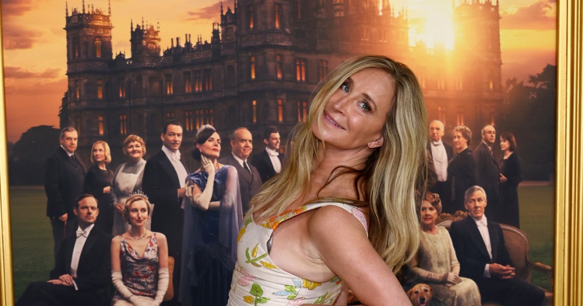 Sonja Morgan in a floral dress, leaning over and doing an over the shoulder pose in front of a Downton Abbey poster