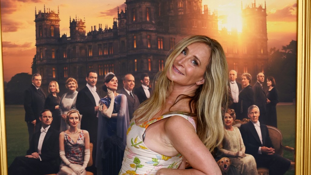 Sonja Morgan in a floral dress, leaning over and doing an over the shoulder pose in front of a Downton Abbey poster