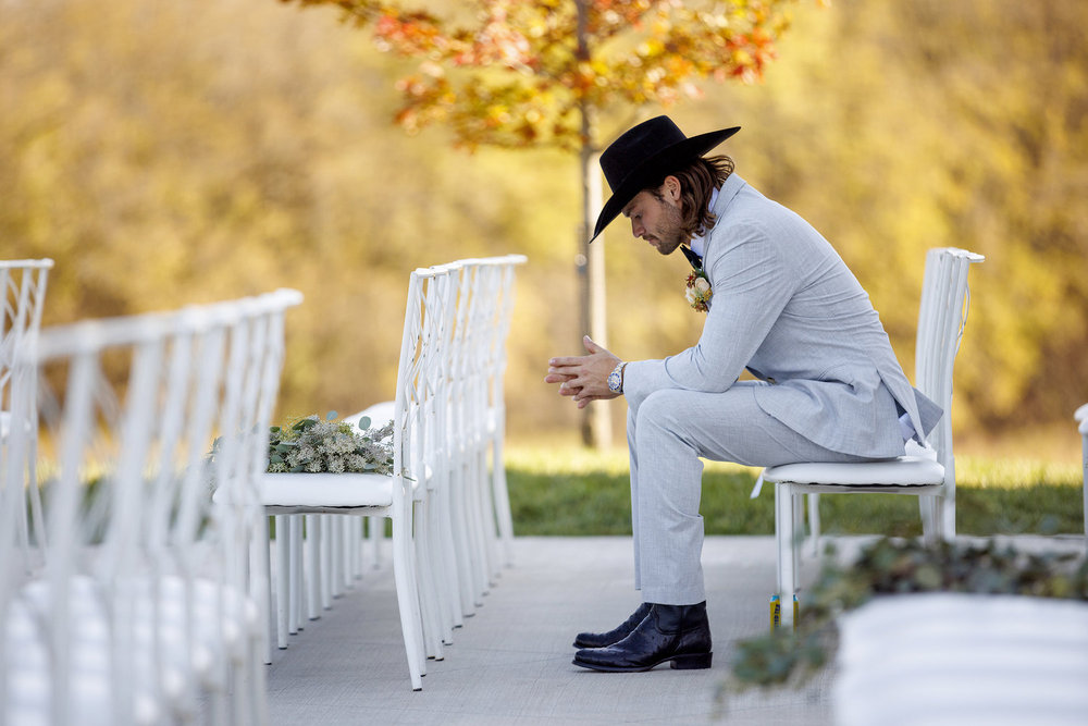 Jesse McBee in a grey suit and black hat, sitting among empty charis at his wedding and looking down at the ground with his hands folded