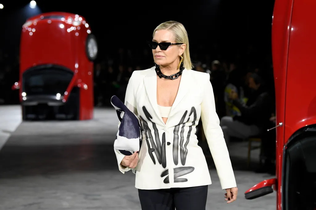 Yolanda Hadid walking in a fashion show in a white blazer, and there are red cards flipped upside down in the background