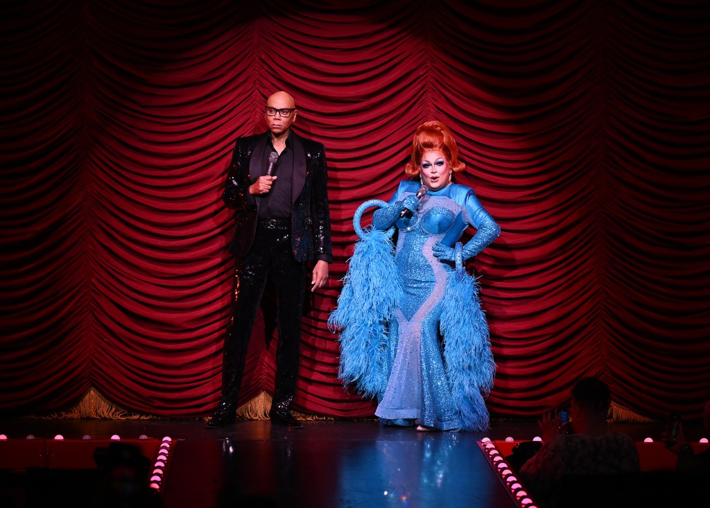 RuPaul in a black suit standing in front of red curtain at Drag Race Live, along with Ginger Minj who is wearing a blue dress