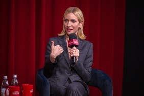 Brittany Snow in a grey suit, sitting in front of a red curtain, holding a microphone and pointing at herself