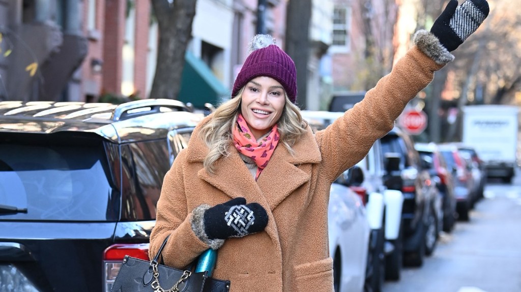 Meghan King in a brown coat and mittens walking down the street and waving