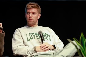 Kyle Cooke with a microphone in his hand, sitting with his legs crossed in a Loverboy sweater