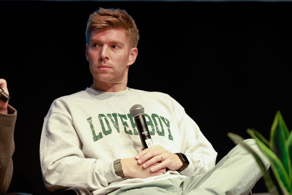 Kyle Cooke with a microphone in his hand, sitting with his legs crossed in a Loverboy sweater