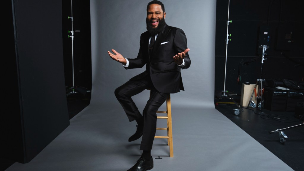 Anthony Anderson sitting on a wooden stool, wearing a black suit