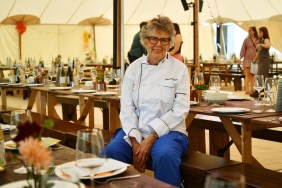 Prue Leith in a chef's jacket and blue pants, sitting on a bench under a large tent