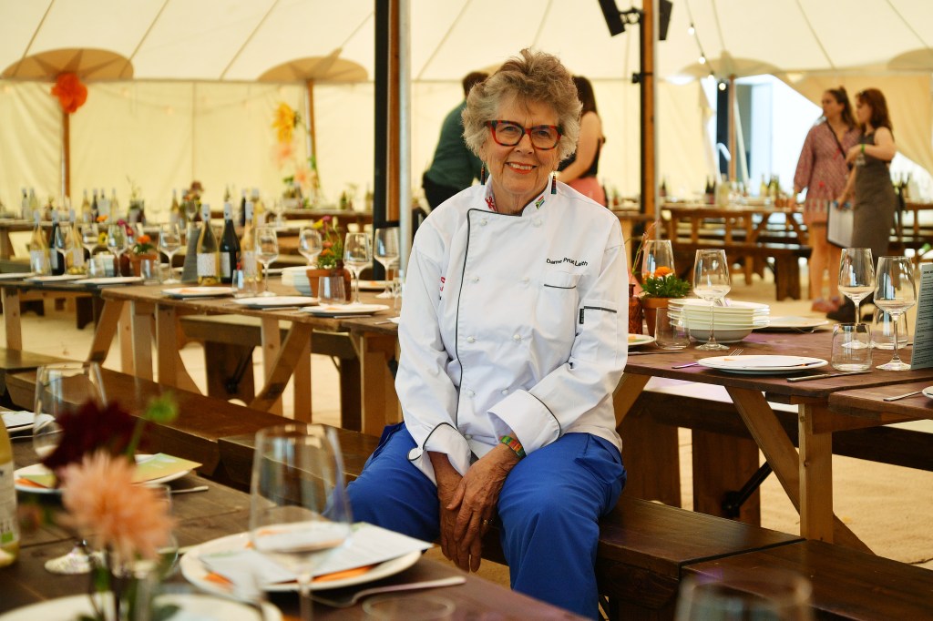 Prue Leith in a chef's jacket and blue pants, sitting on a bench under a large tent