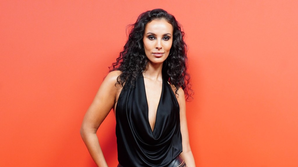 Maya Jama in a black dress staying against an orange backdrop