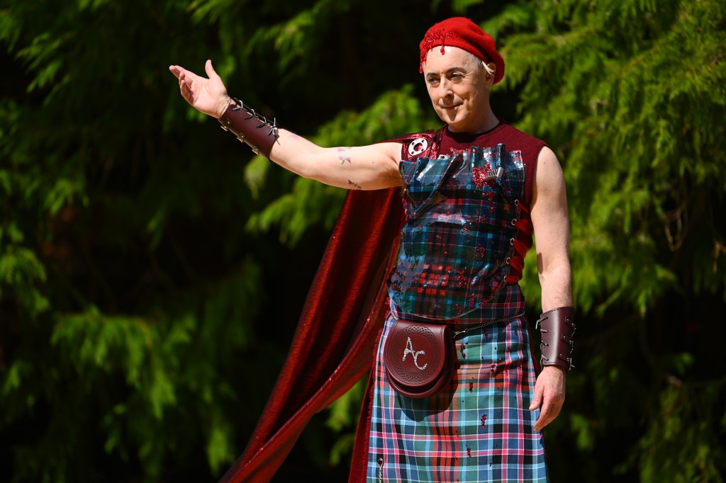 Alan Cumming dressed in red presenting a challenge on The Traitors