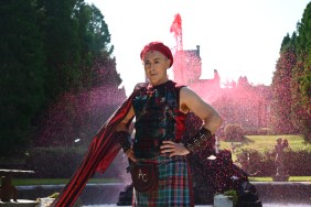 Alan Cumming with his hands on his hips, wearing a kilt, standing in front of a red fountain