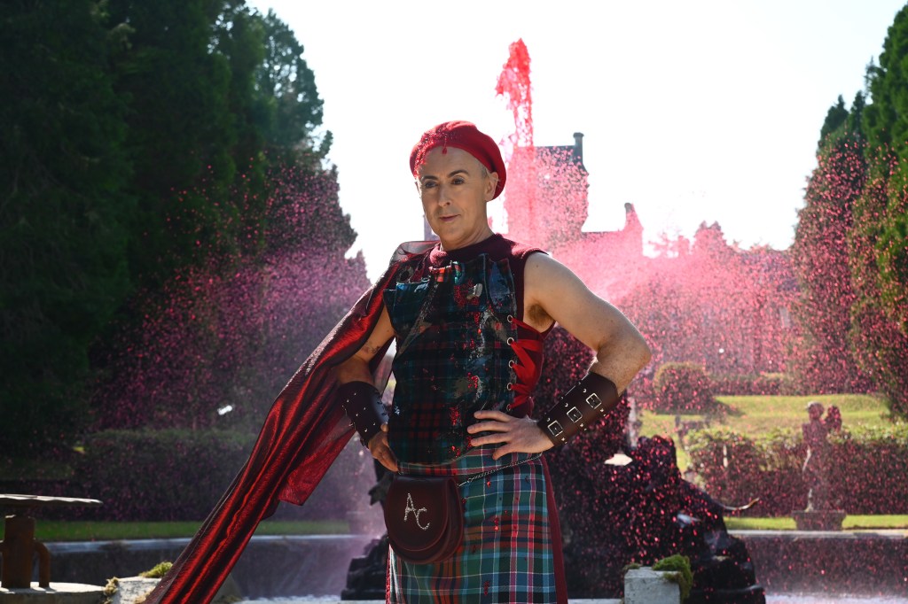 Alan Cumming with his hands on his hips, wearing a kilt, standing in front of a red fountain