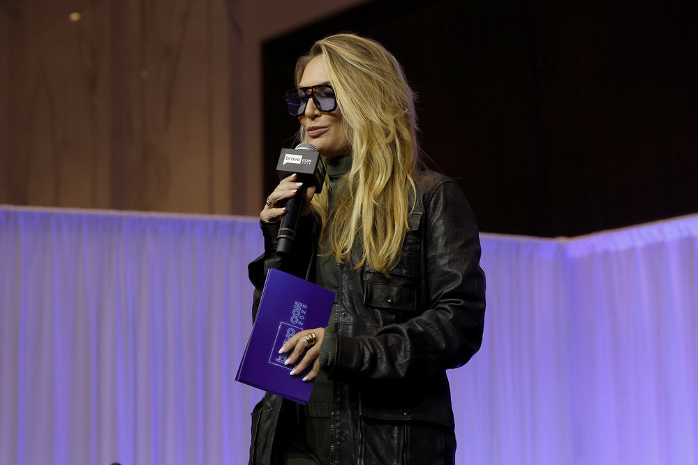 Kate Chastain on stage in a black coat, holding a microphone at BravoCon 2025