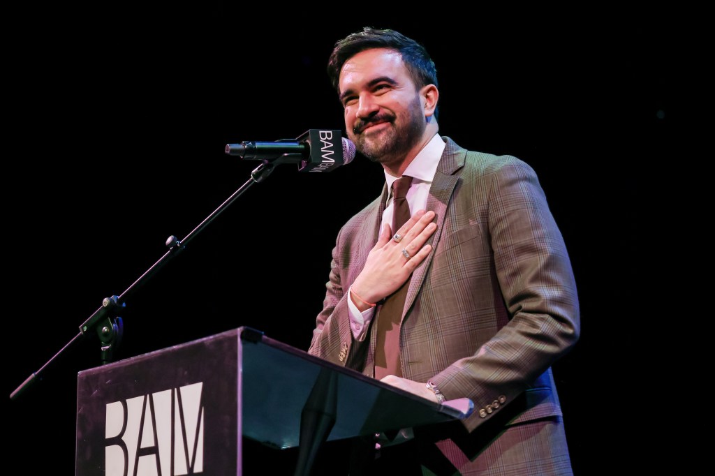 Mayor Zohran Mamdani in a brown suit speaking at a podium with his hand over his heart
