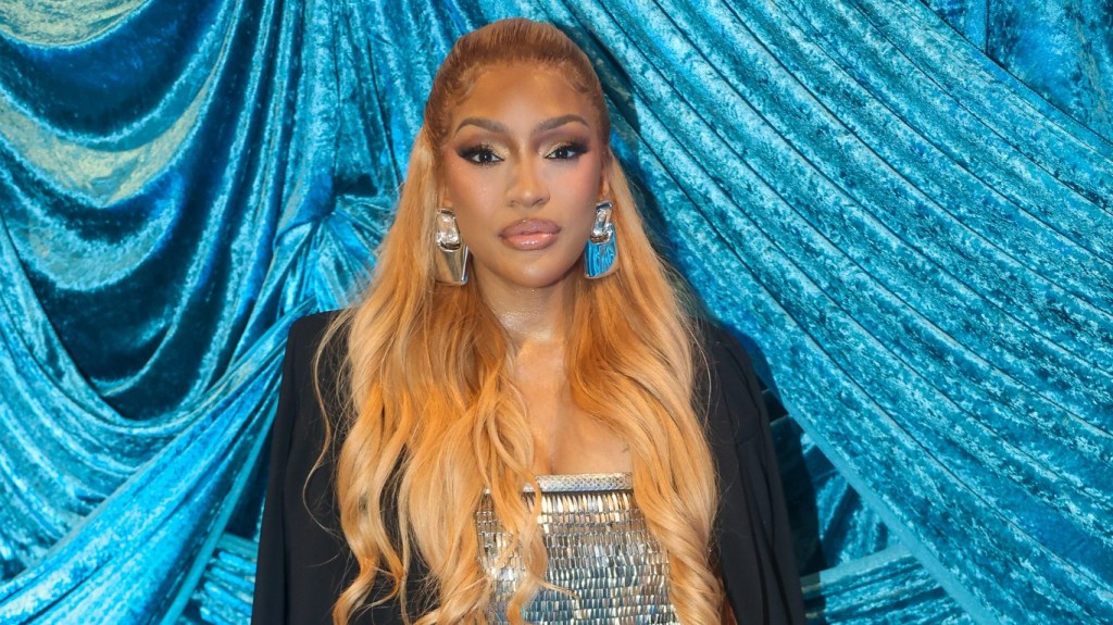 Drew Sidora in a silver top and black blazer posing in front of blue drapery