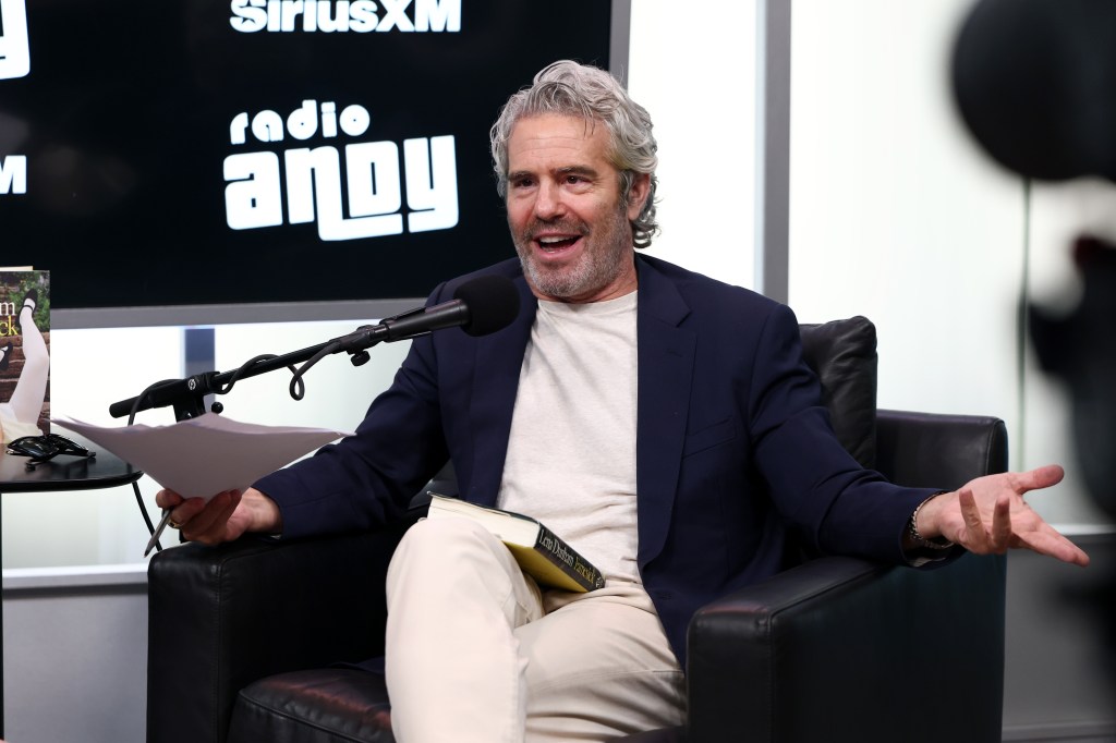 Andy Cohen with a book in his lap, holding his arms out in disbelief while recording for Radio Andy