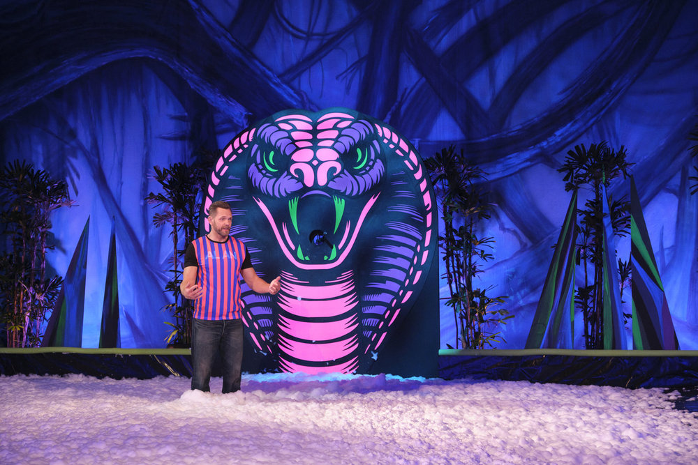Joel McHale in a foam pit in front of a neon colored snake on House of Villains