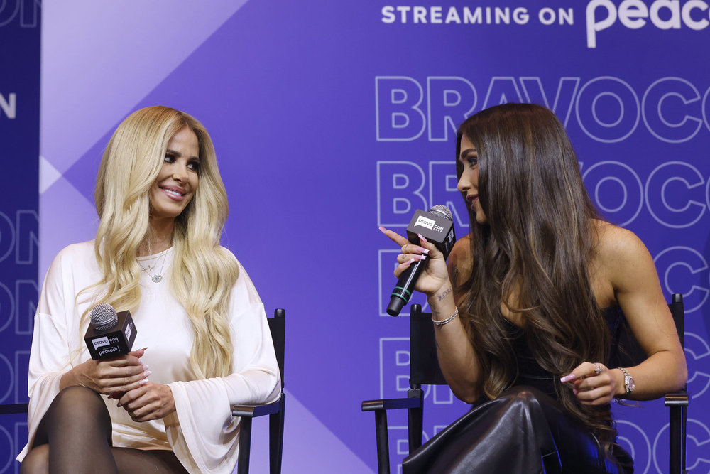 Kim Zolciak wearing white on stage at BravoCon talking to Ariana Madix who is wearing a black dress and pointing at Kim